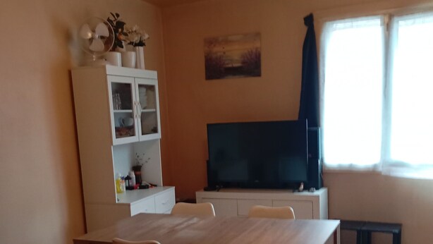 Un salon comprenant une table à manger en bois, quatre chaises, un téléviseur posé sur un meuble, un buffet aux portes vitrées et un tableau accroché au mur.