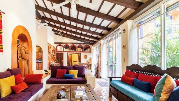 Amplia sala de estar con techo de madera, un conjunto de sofás con cojines coloridos, una mesa de centro con libros y objetos decorativos, y ventanas grandes con vistas a la vegetación.