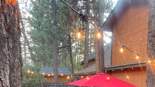 An outdoor patio with wicker furniture, a red umbrella, and string lights. - Pet friendly cabin in the woods perfect for your adventures (Big Bear)