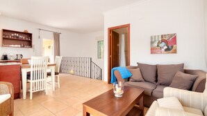 Una sala de estar con un sofá, una mesa de centro de madera, un comedor con sillas, una zona de cocina con estantes y vistas al exterior.