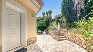Ein pflasterbelagter Eingangsbereich mit einer weißen Tür, eine Steintreppe und ein Garten mit verschiedenen Pflanzen und einer Palme.