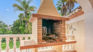 Ein Outdoor-Ziegelofen-Grill auf einem Balkon mit weißer Reling, einer weißen Wand und Blick auf Palmen sowie ein Haus im Hintergrund.