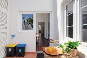 Une façade propre et blanche, avec une fenêtre, une porte donnant sur un couloir et un petit espace extérieur aménagé avec une table et des chaises.