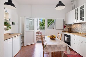 A compact kitchen with white cabinets, a dining table with a patterned tablecloth, and a view through an open door to a balcony.