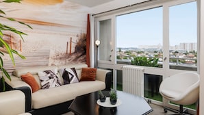 Un salon moderne avec un canapé, une table basse et un fauteuil. Une grande fenêtre offre une vue sur les bâtiments et la verdure.