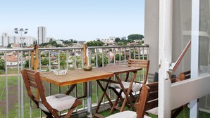 Un balcon avec une table et des chaises en bois, donnant sur un paysage urbain composé d’immeubles et d’espaces verts.