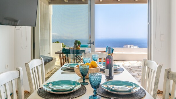 A dining table with blue glassware, plates, and a bottle, set against a backdrop of a scenic ocean view. - Country House 'Allegro' with Sea View, Wi-Fi and Air Conditioning (Tinos)