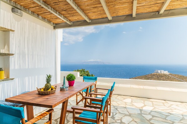 A wooden table with chairs and a basket of fruit on a terrace with a view of the sea.