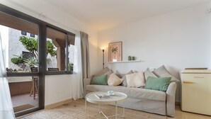 Una sala de estar con sofá, una mesa redonda pequeña, un refrigerador y una ventana con cortinas.