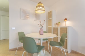 A dining area with a round table, four chairs, a hanging light fixture, a vase with flowers, and a wall-mounted cabinet with a bowl and a lamp. - Oasis Duna - Casa Amanay: Shared Pool, Private Terrace & Wi-Fi for Your Vacation (Corralejo)