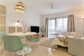 A modern living room with a round dining table, green chairs, a sofa, a TV, and a large window with curtains. - Oasis Duna - Casa Amanay: Shared Pool, Private Terrace & Wi-Fi for Your Vacation (Corralejo)
