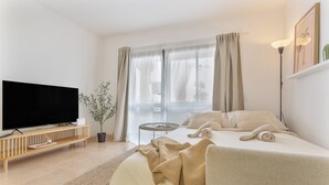 Una habitación de hotel moderna con una televisión de pantalla plana, una planta en maceta, una cama con ropa de cama color beige, una mesita de noche y un ventanal con cortinas.