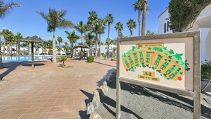 Un patio con baldosas, palmeras, una piscina y un cartel con información.