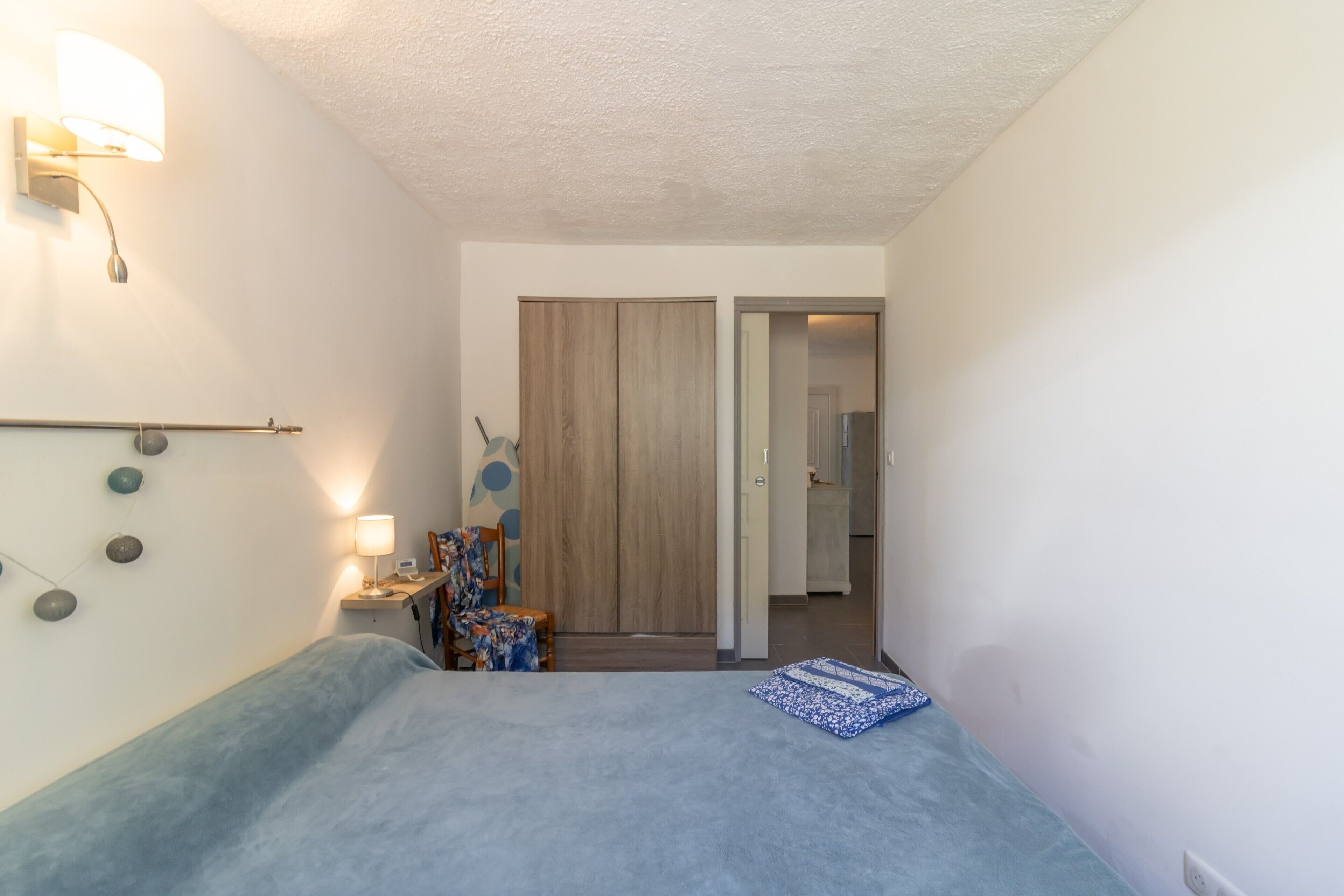 Un dormitorio con cama, mesita de noche, lámpara, silla, armario y puerta que da a otra habitación.