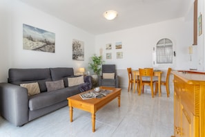 A modern living room with a grey sofa, wooden coffee table, and a dining area with wooden chairs and a table. - Pool-View Bungalow with Air Con & Private Roof Terrace, Central Corralejo (Corralejo)