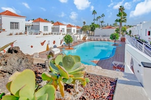 A pool area with a clear blue pool, white buildings with red roofs, and a small tree in the foreground. - Pool-View Bungalow with Air Con & Private Roof Terrace, Central Corralejo (Corralejo)