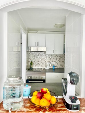 Una cocina con armarios blancos, salpicadero de azulejos, una tetera, una cafetera y un frutero sobre la encimera.