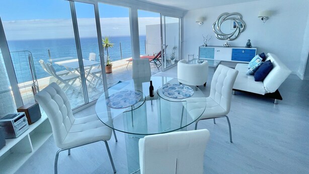 Ein modernes Wohnzimmer mit Glastisch, weißen Stühlen, einer Couch und Blick auf den Ozean.