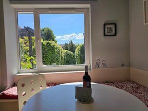 A room with a window overlooking greenery, a table with a bottle, and a chair.