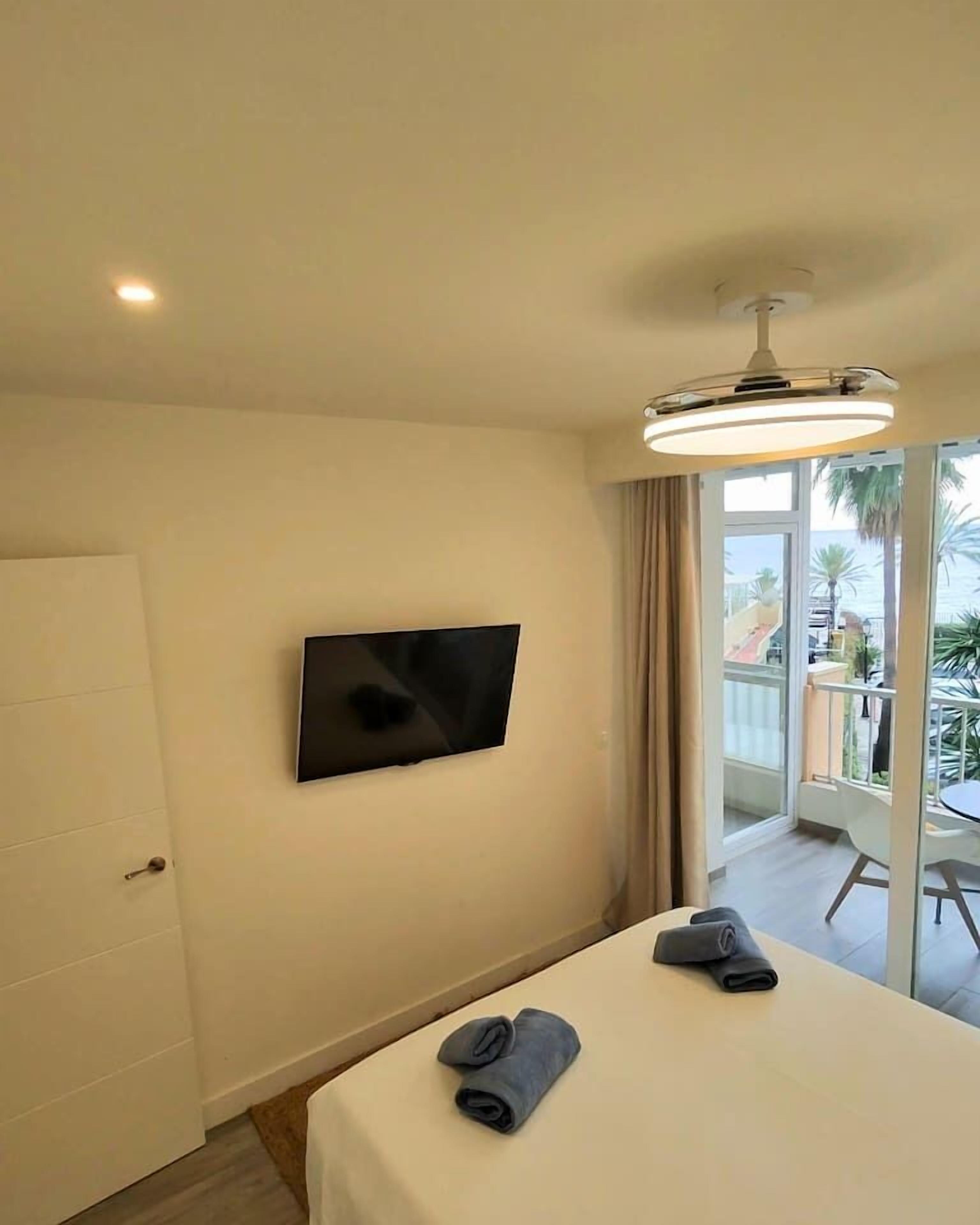 Habitación de hotel con cama, televisor montado en la pared y balcón con vistas a palmeras.