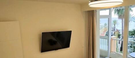 Habitación de hotel con cama, televisor montado en la pared y balcón con vistas a palmeras.