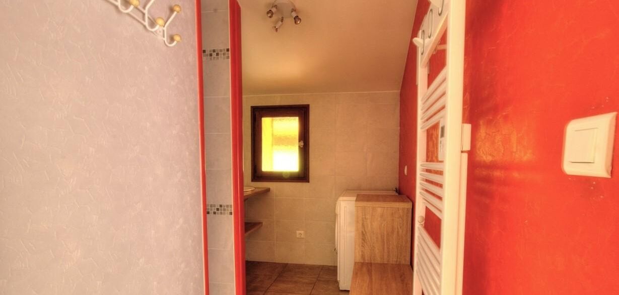 Ein schmaler Flur führt zu einem Badezimmer mit Fenster, roter Wand und weißer Tür.