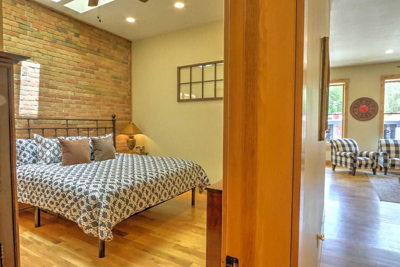 A bedroom with a brick wall, a bed with patterned bedding, a nightstand with a lamp, and a view of the living area with checkered chairs.