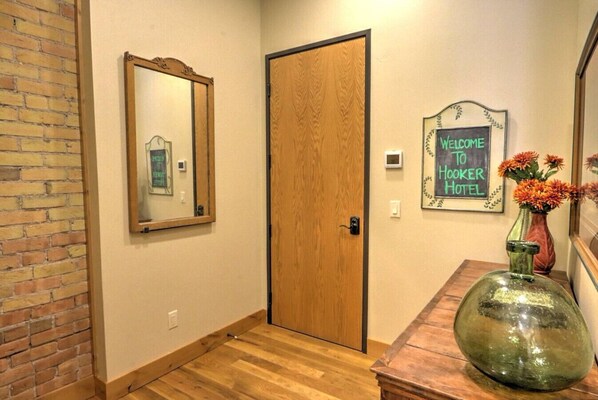 Un pasillo con una puerta de madera, un espejo, un cartel de bienvenida y un jarrón con flores sobre una mesa.