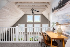Una stanza con soffitto spiovente, una ventola a soffitto, un tavolo da pranzo con sedie, una ringhiera bianca e vista sul verde attraverso le finestre.