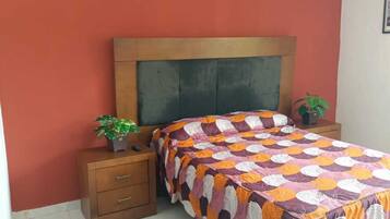 A bedroom with a wooden headboard, a colorful quilt, a bedside plant, and a small wooden nightstand.