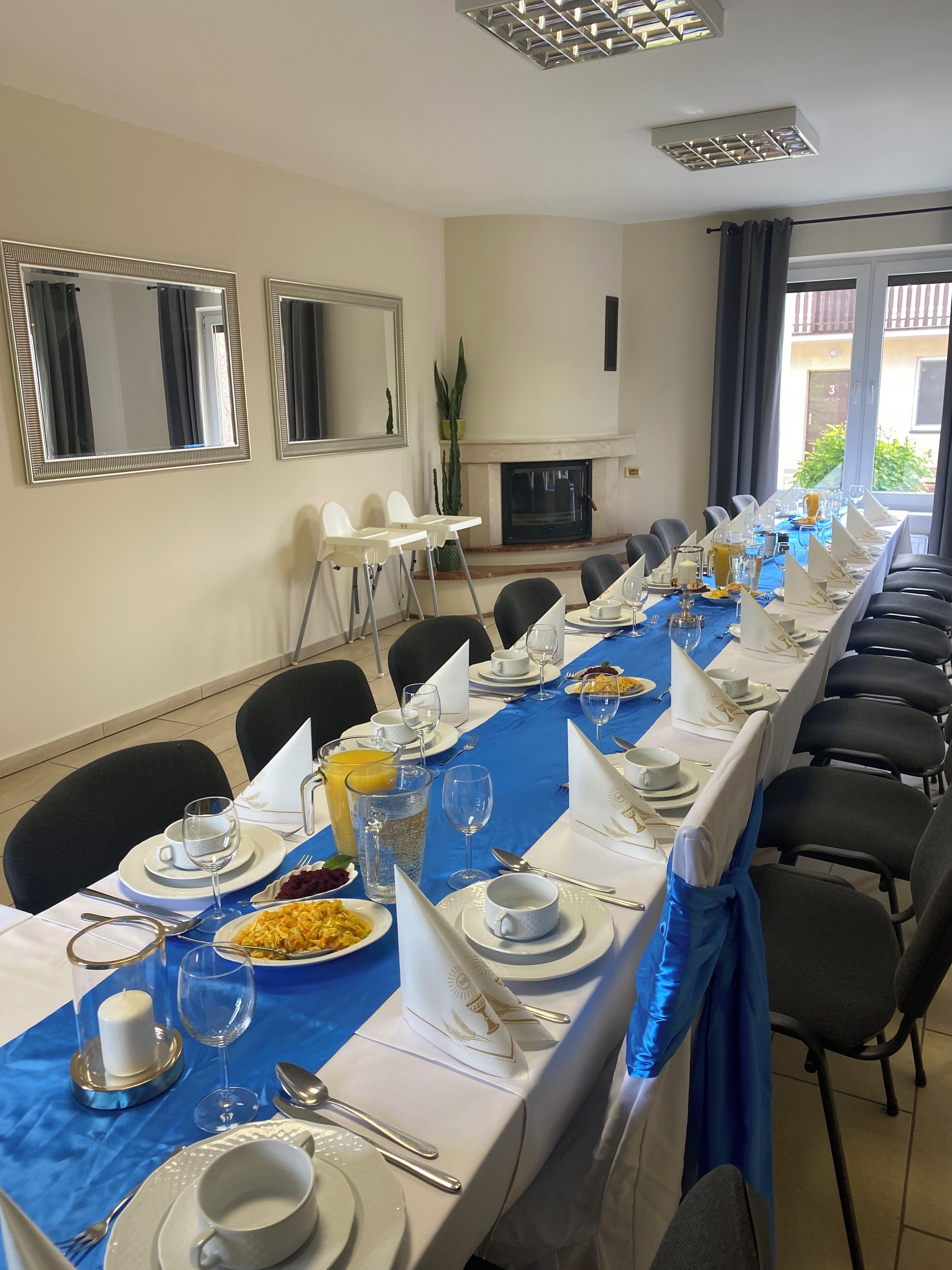 Une longue table à manger, dressée avec des assiettes, des verres et des couverts, entourée de chaises et recouverte d’une nappe bleue.