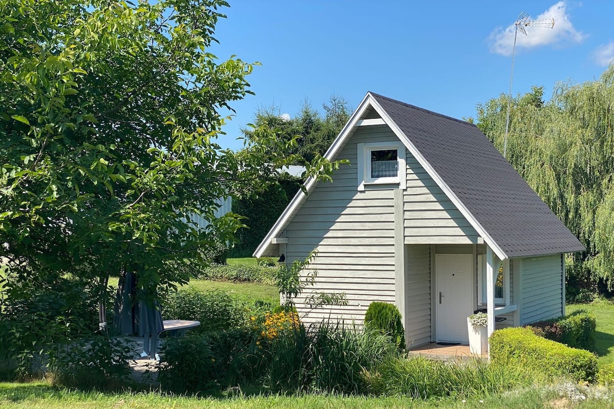A small, single-story house with a gabled roof, surrounded by lush greenery and a clear blue sky.