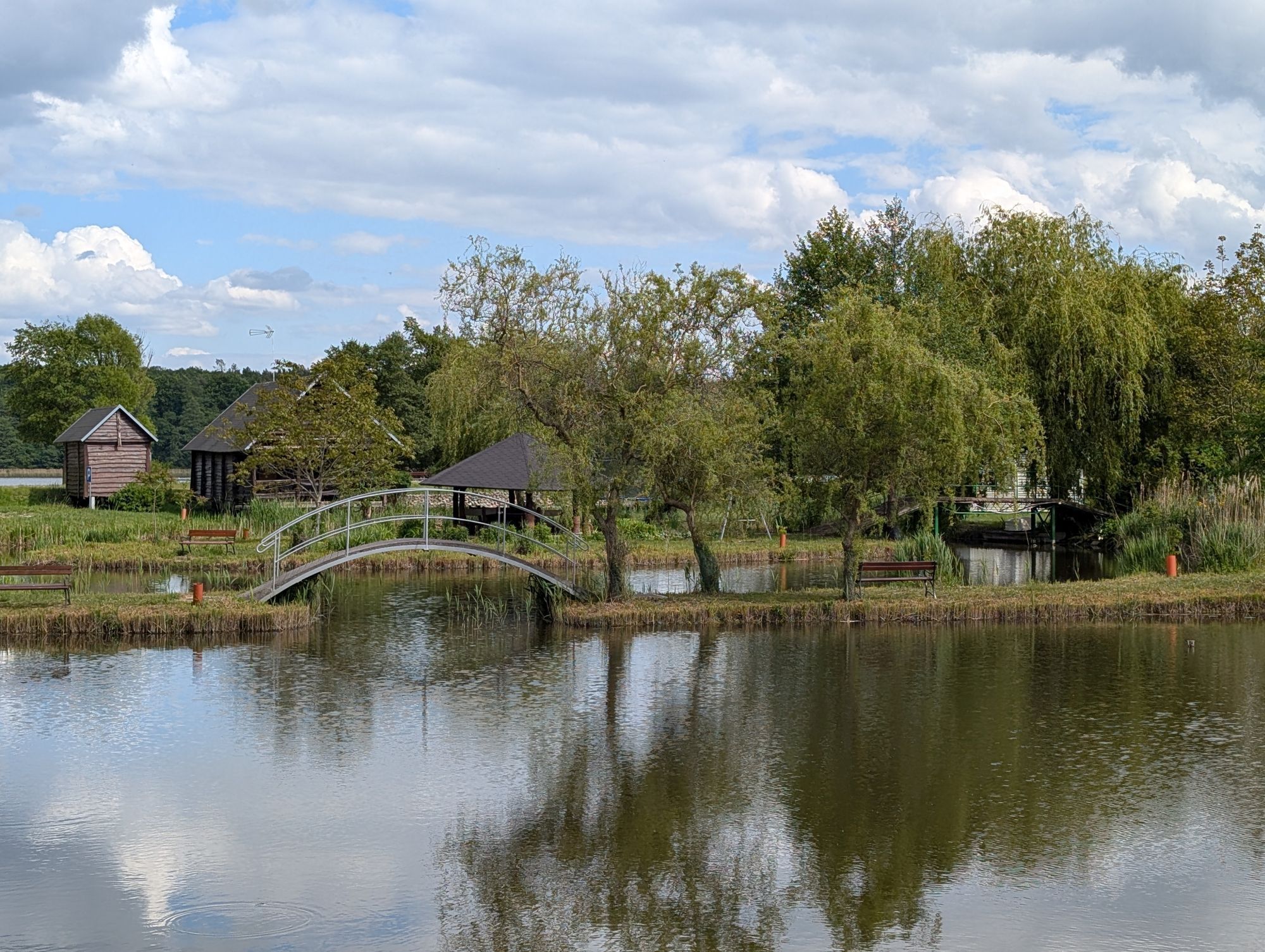 Uma paisagem tranquila à beira do lago com uma ponte de madeira, um gazebo e uma cabana pequena.