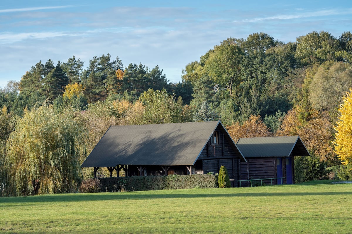 A rustic cabin with a sloped roof, surrounded by a grassy field and dense trees with autumn foliage.