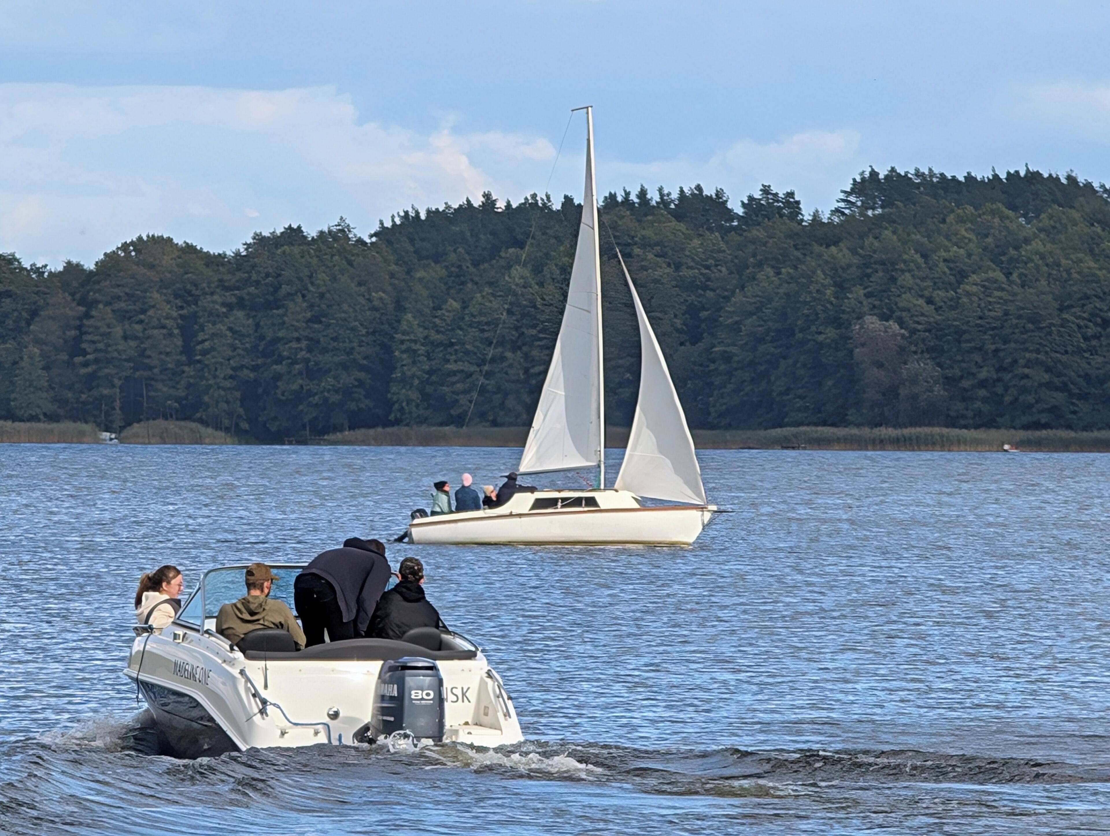 A boat with people on it, a sailboat in the background, and a forested shoreline.