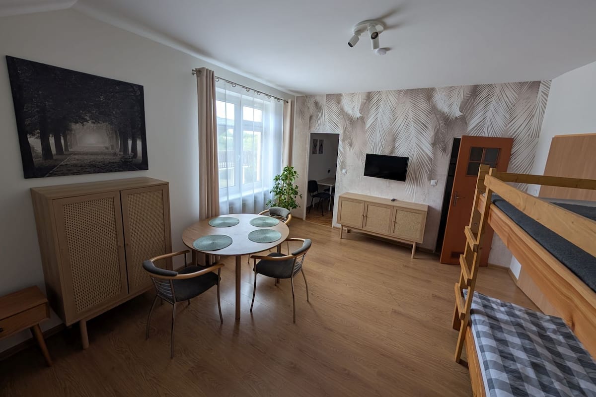 A room with a round table, chairs, a TV mounted on the wall, a bunk bed, and a large window with curtains.