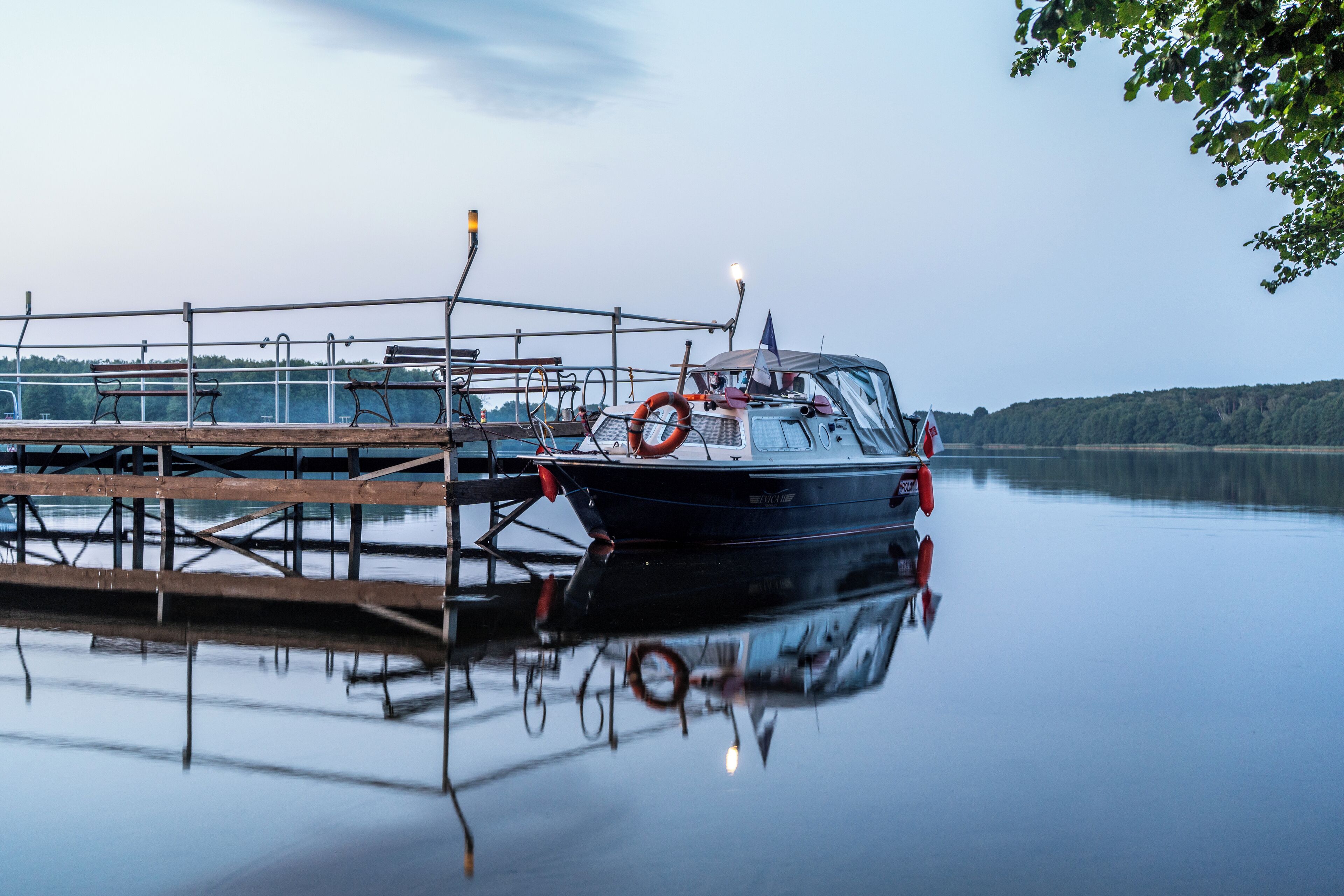 A boat docked at a pier with a lifebuoy attached, surrounded by calm water and a forested landscape in the background.