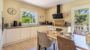 A modern kitchen with a dining area, featuring a clock, a coffee maker, and a view of greenery outside. - Villa de L'Uba - Welkeys (Toulon)