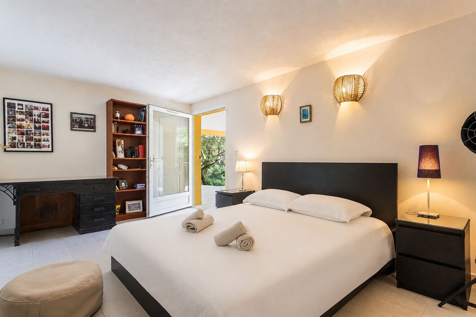 Habitación de hotel con cama, mesitas de noche, escritorio, estantería y puerta corrediza de vidrio al exterior.