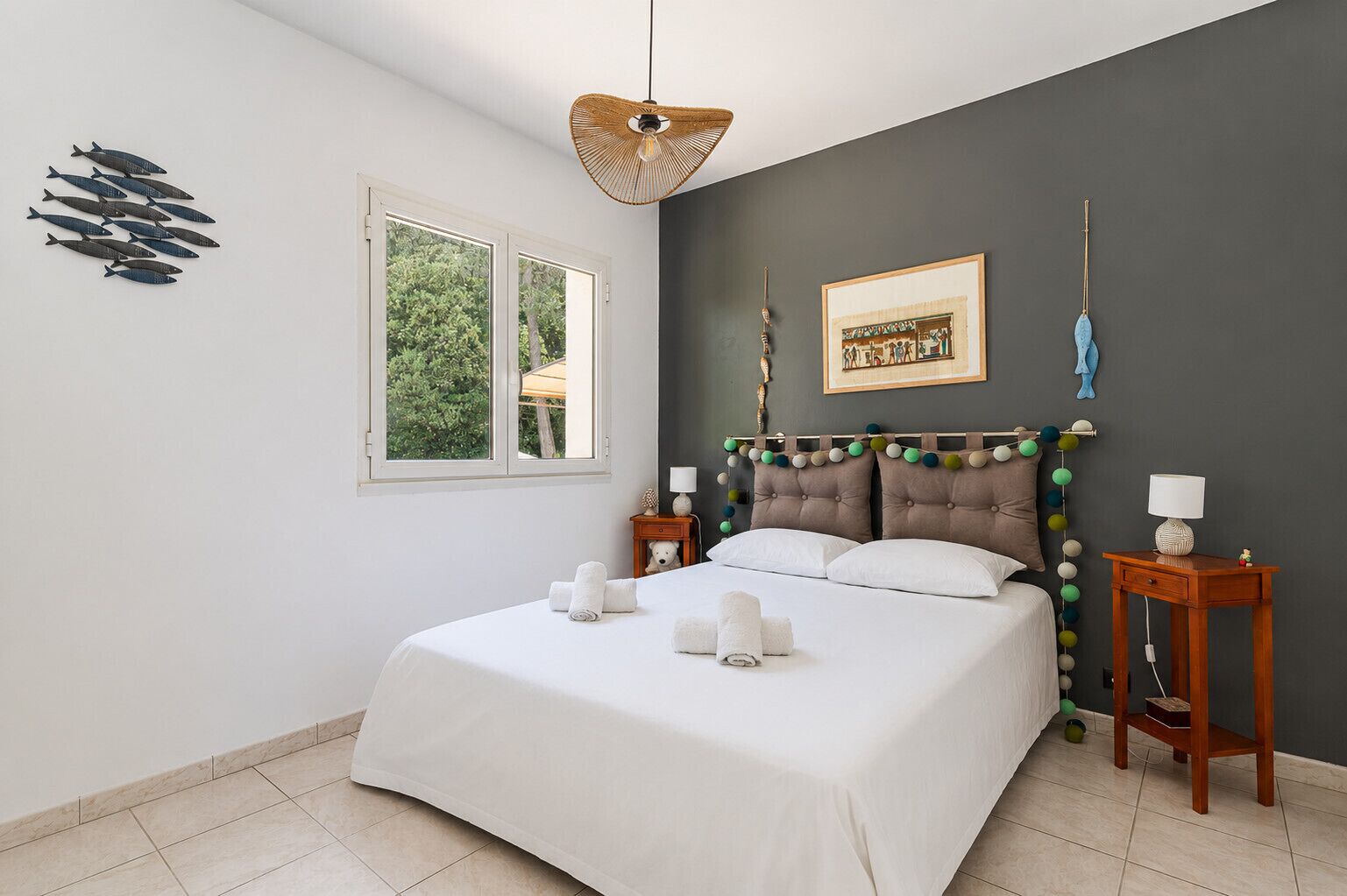 Une chambre à coucher comprenant un lit, des tables de chevet, un luminaire suspendu, une fenêtre et un élément décoratif mural.