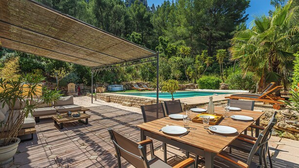 An outdoor patio with a wooden table set for dining, surrounded by chairs, and a pool in the background. - Villa de L'Uba - Welkeys (Toulon)