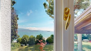 Blick durch ein Fenster mit einer Tür auf eine grüne Fläche mit Bäumen und einem Gewässer.