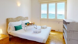 Ein Schlafzimmer mit einem Bett, einem Nachttisch, einem Fenster mit Meerblick und einer Holzkommode.
