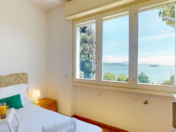 A bedroom with a bed, a nightstand, a lamp, and a window with a view of the sea and mountains. - Villa 7 Bello by Bookinggardalake (Manerba del Garda)