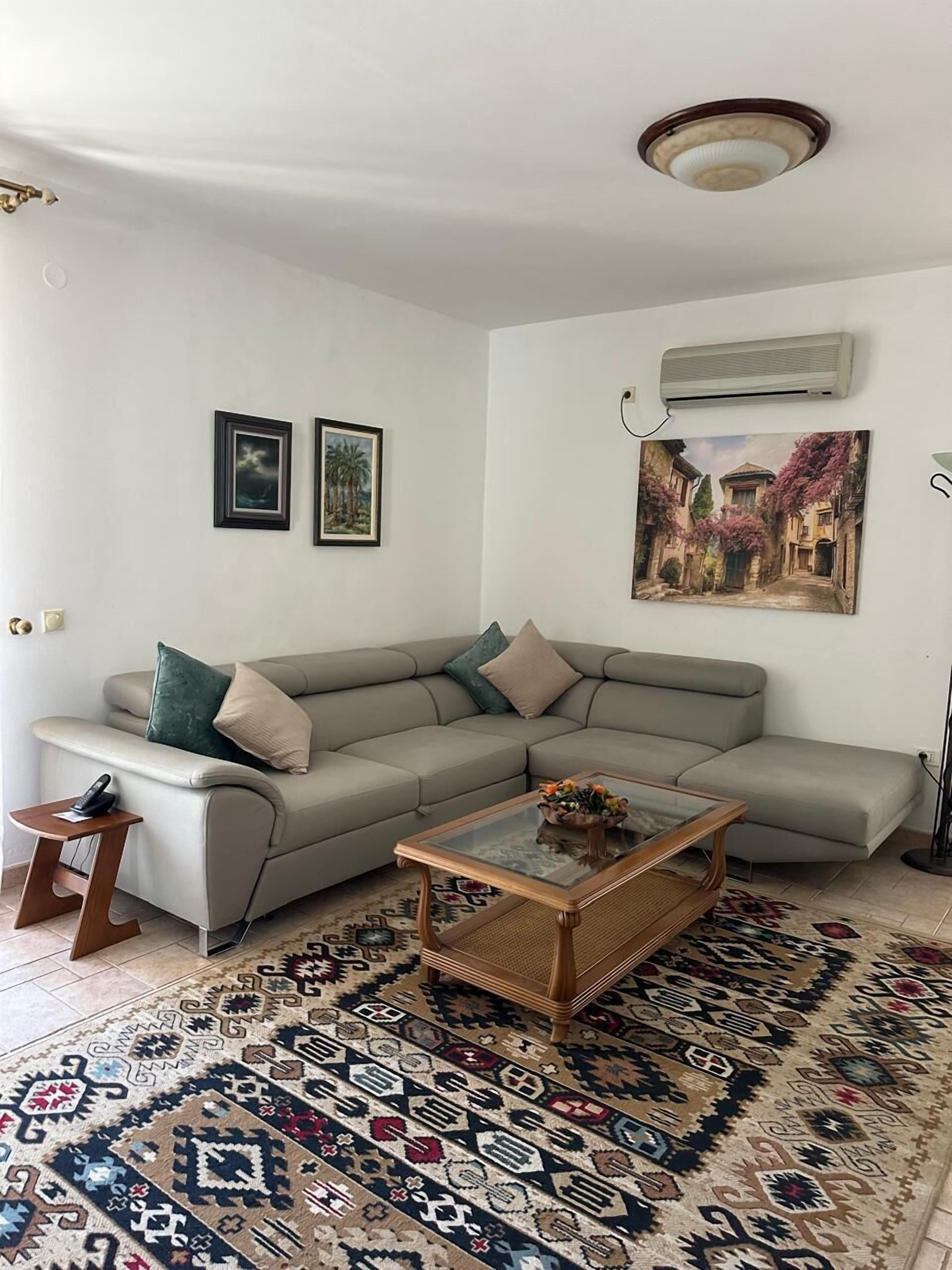 Una sala de estar con sofá modular, una mesa de centro de vidrio, alfombra estampada, aire acondicionado de pared y obras de arte enmarcadas.