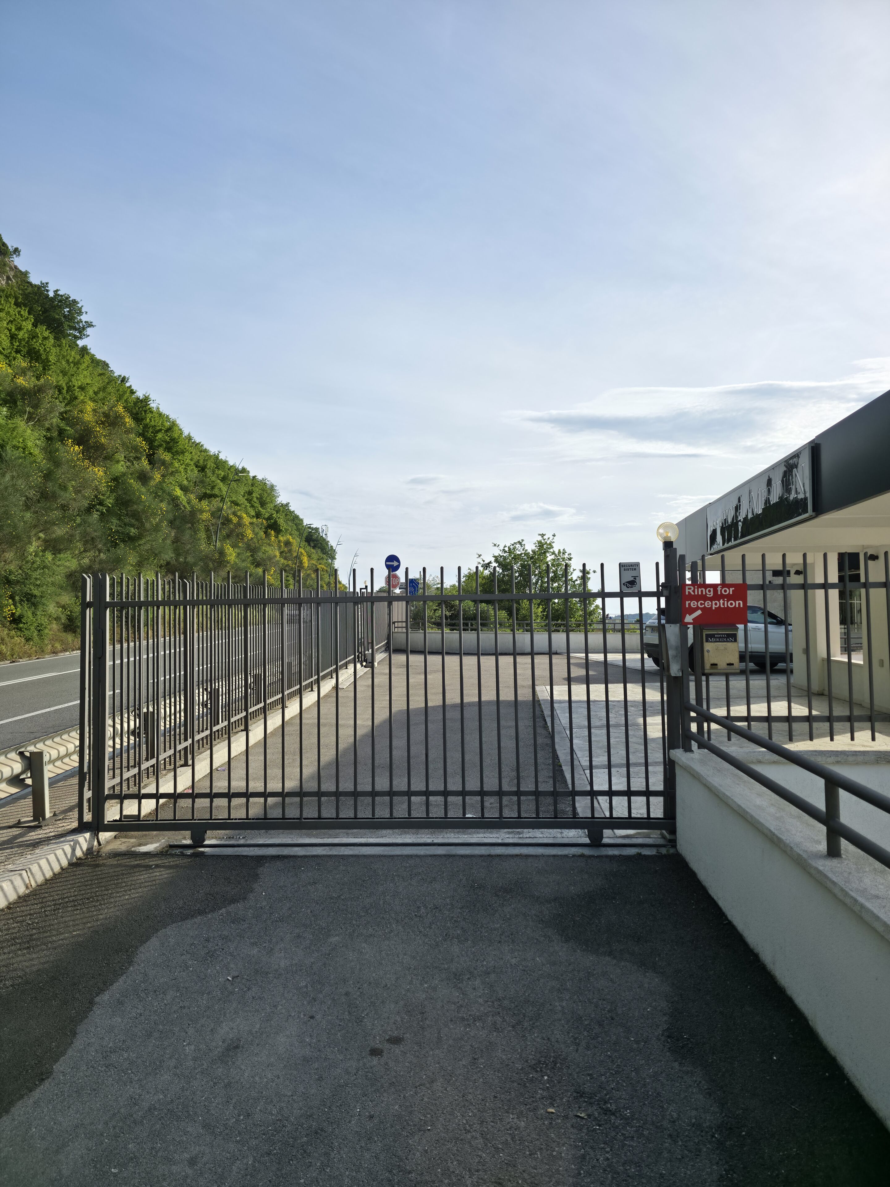 Une entrée fermée par un portail mène à un bâtiment, avec un ciel dégagé et de la verdure en arrière-plan.