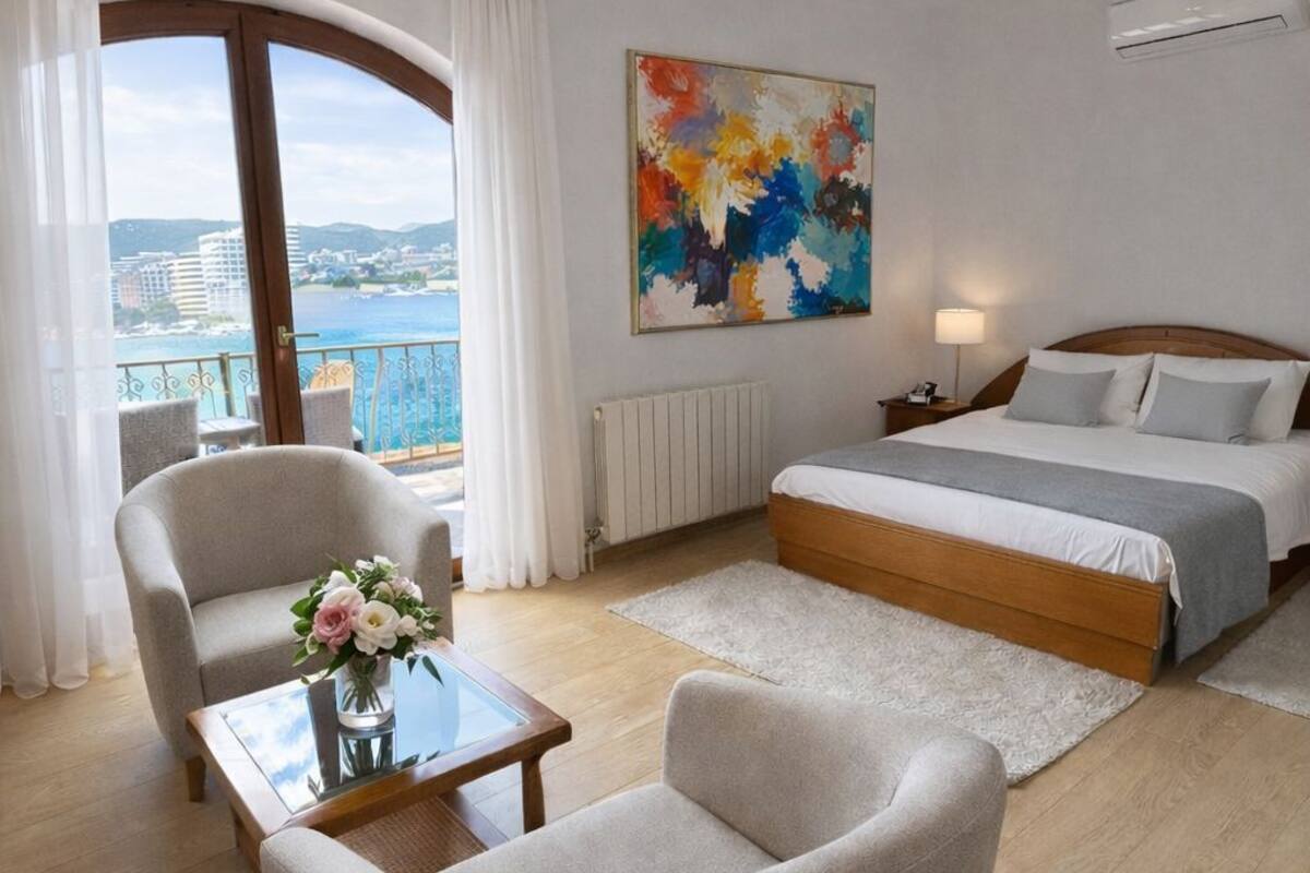 Quarto de hotel moderno com uma cama, uma cadeira, uma mesa com um vaso de flores, um quadro na parede e vista para a cidade pela janela.