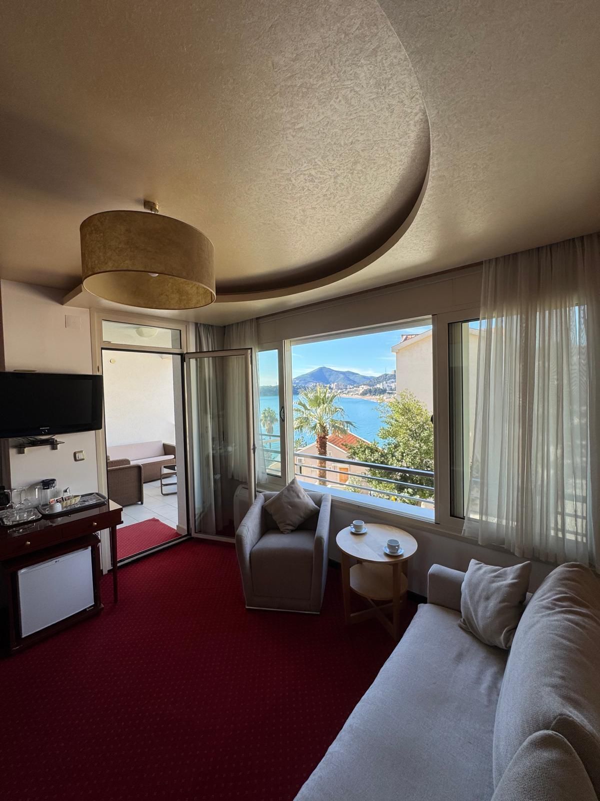 A hotel room with a bed, a TV, a desk with a coffee maker, a chair, a small table with a cup, and a balcony with a view of the sea and mountains.