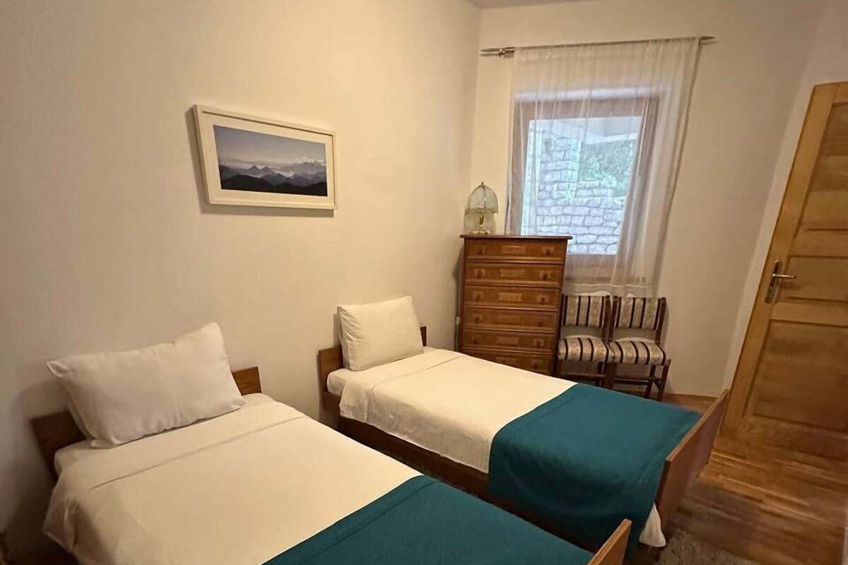 Quarto com duas camas, cômoda, cadeira, janela com cortinas e porta.
