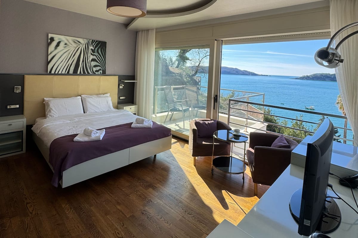 Quarto de hotel com cama, escrivaninha com computador, cadeira, mesa e vista para o mar.
